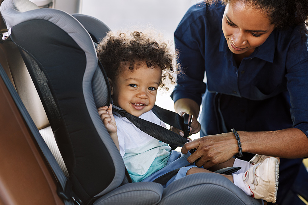 happy little boy looking at camera while his mother buckles him into a car seat